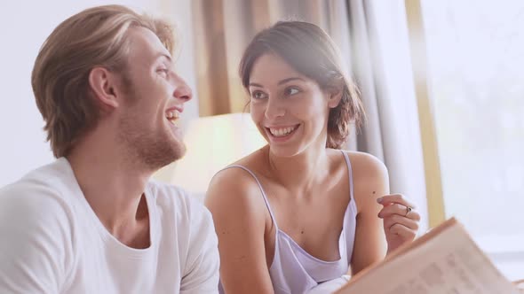 Beautiful Caucasian Pair Sitting at Bed Talking with Each Other Smiling Laughing Reading Magazine in