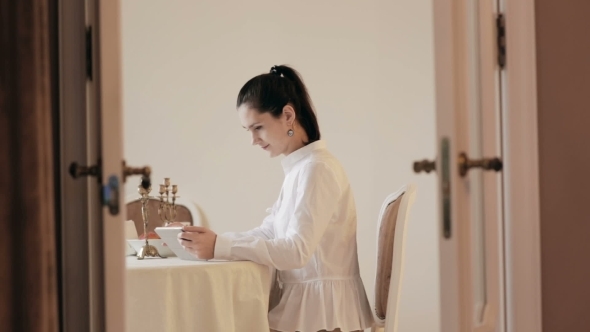 Woman Using Tablet And Drinking Coffee By Table 