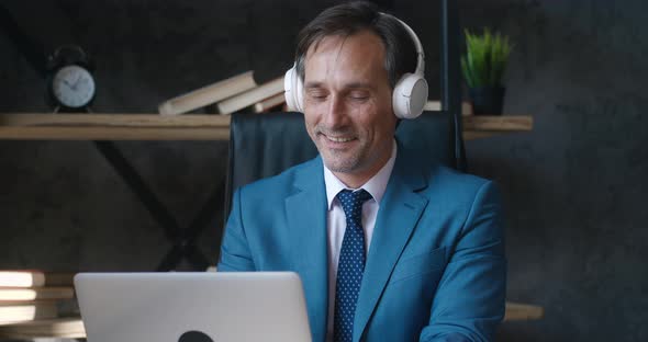 Focused Smiling Mature Male Entrepreneur with Headphones Listening Music and Typing on Laptop