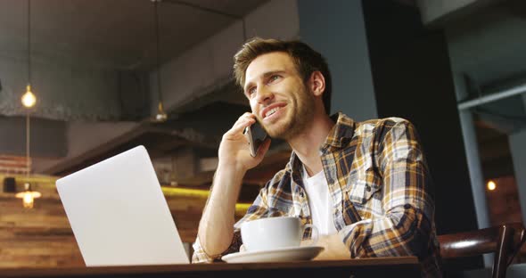 Man talking on mobile phone while using laptop in cafe 