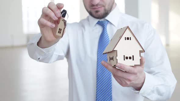 Realtor Man is Holding Keys with Keychain Form of House and Wooden Figure of House Front View