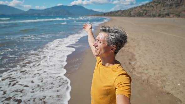 Happy Middle Aged Greyhead Man Stands at Sandy Sea Coast with Raised Hands