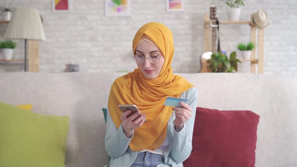 Positive Young Muslim Woman in Hijab Smiling Holding Smartphone and Bank Card Sitting on Sofa in