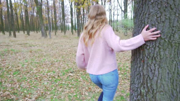 Attractive Blonde Girl Hiding in the Forest and Leaning Against the Old Tree