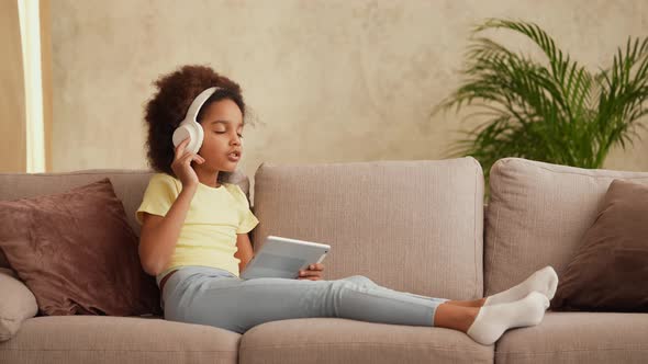 Portrait Little African American Girl in Big White Headphones Listens Music Using Digital Tablet