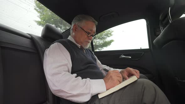 4K Senior businessman sitting on car back seat and writing business plan in the book