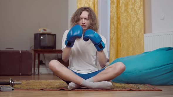 Young Skinny Man with Long Curly Hair Sitting on Retro Carpet in Boxing Gloves Fighting Shadow and
