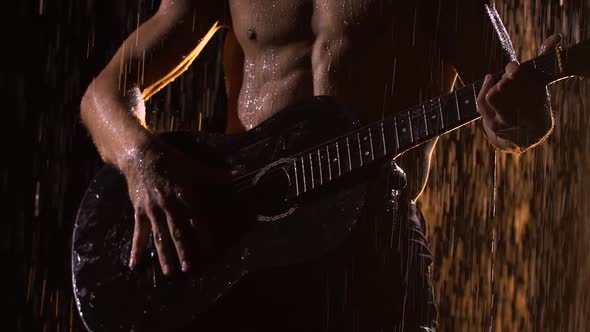 A Man with a Naked Torso Wet From Raindrops Plays the Guitar While Sitting on the Surface of the