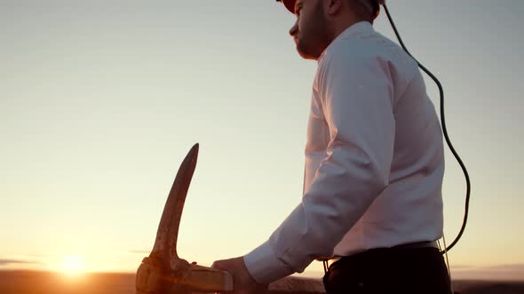 Man stands background sunset in helmet and throws pickaxe over shoulder, side view. Businessman