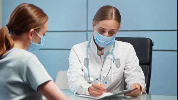 Lady general practitioner in disposable face mask and medical coat talks to blonde hospital visitor