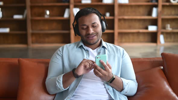 Cheerful Indian Guy Using Smartphone and Wireless Headphones for Listening Music