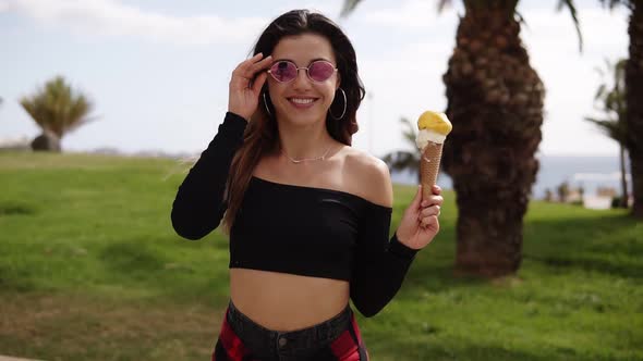 Beautiful Woman in Sunglasses Walking in the Park with Palm Trees Holding Yellow Cone Ice Cream