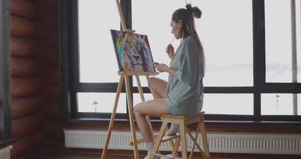 Calm Young Woman Dressed Pajamas is Drawing Picture on Easel Near Window at Home Holding Paintbrush