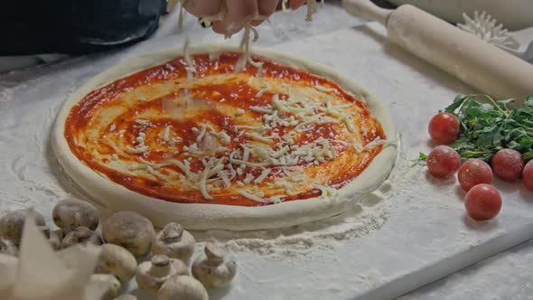 Professional Chef Pours Grated Cheese Onto a Pizza Base with Tomato Paste on the Table Decorated