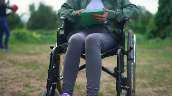 Unrecognizable Girl in Wheelchair with Tablet Resting in Summer Park After Accident