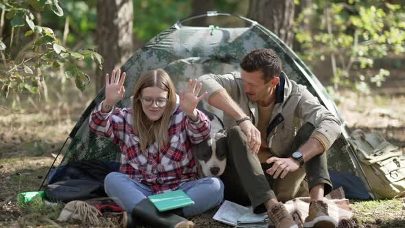 Front View Cheerful Woman and Man Laughing Sitting at Tent with Dog in Forest