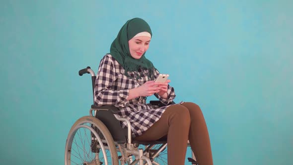 Young Muslim Woman Disabled Person in a Wheelchair Uses the Phone on a Blue Background