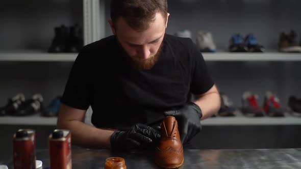 Portrait of shoemaker in black latex gloves applying paint on parts of light brown leather shoes.