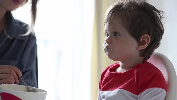 mother feeds a little toddler boy with a spoon during lunch in the room.