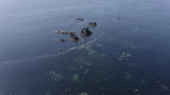 Aerial descends to shallow sea water near jagged ocean rocks
