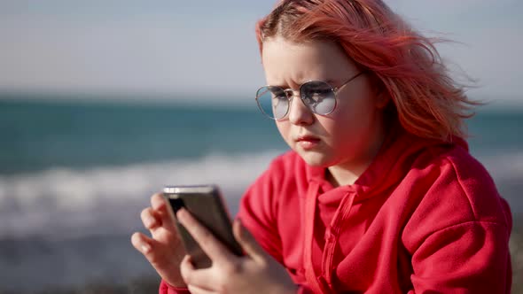 A Serious Teenage Girl with Pink Hair is Looking Through a Mobile Phone Sitting on the Ocean