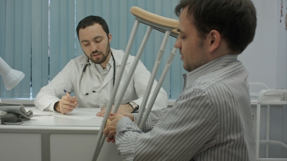 Male Bearded Doctor In Clinic Consult Sick Client With Crutches.