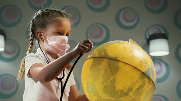 Little Girl Wearing a Medical Mask and Listening to a Globe