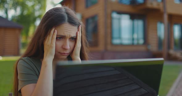 Frustrated Frowning Young Girl Looks at Laptop Monitor Sitting at Table in Courtyard of Country