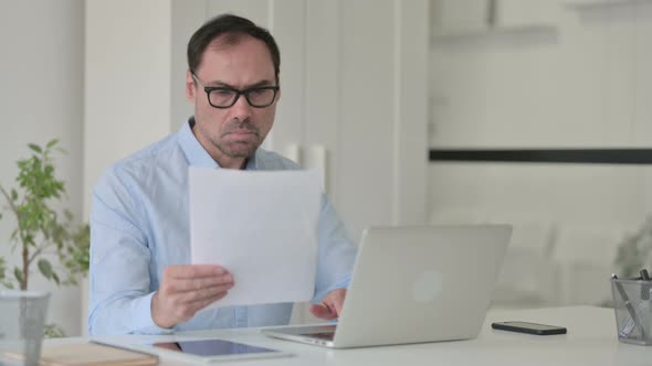 Middle Aged Man Reading Documents While Using Laptop