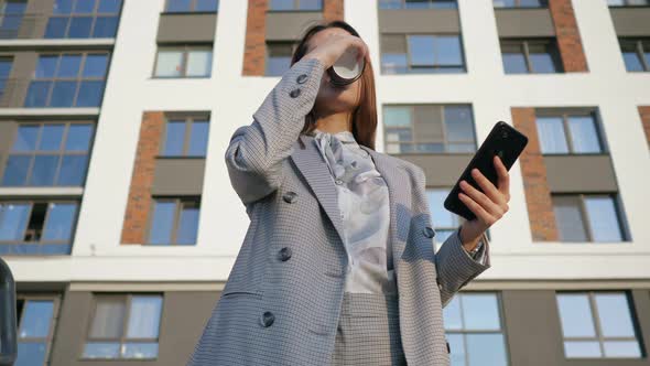 Young Woman in a Business Suit Looks at the Phone and Drinks Coffee From a Plastic Cup on the