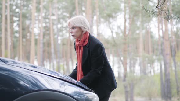 Portrait of Serious Beautiful Blond Woman Opening Hood of Broken Car Outdoors. Young Caucasian Lady