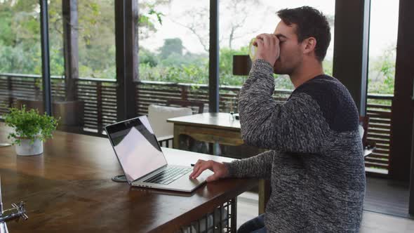 Caucasian man working at home, using laptop and drinking coffee in dining room