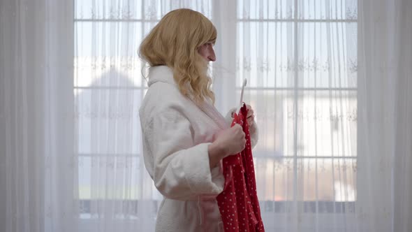 Side View Happy Travesty Woman in Bathrobe Admiring Red Dotted Dress Standing in Bedroom at Home