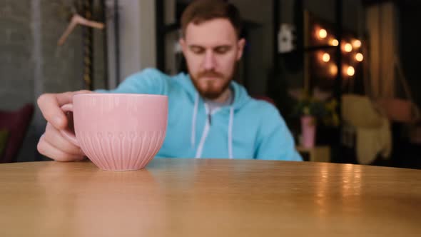 Bearded Man Guy in Blue Sweater Hoodie Sits at a Table in a Cafe Looks at the Phone and Drinks