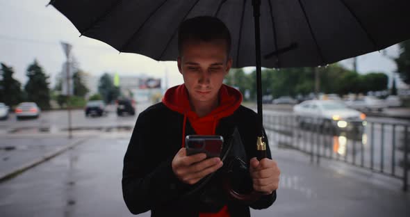 Man Walking in City Under Umbrella Using Smartphone