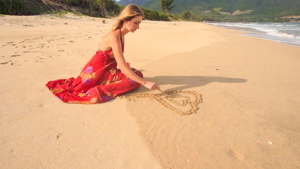 Woman in Red Dress Sits Draws Heart on Sand of Wet Beach