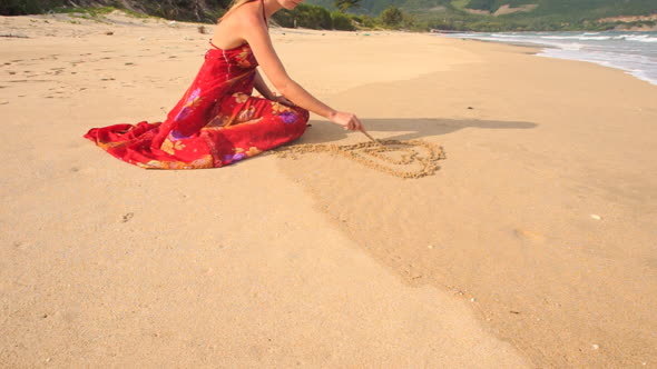 Woman in Red Dress Sits Draws Heart on Sand of Wet Beach