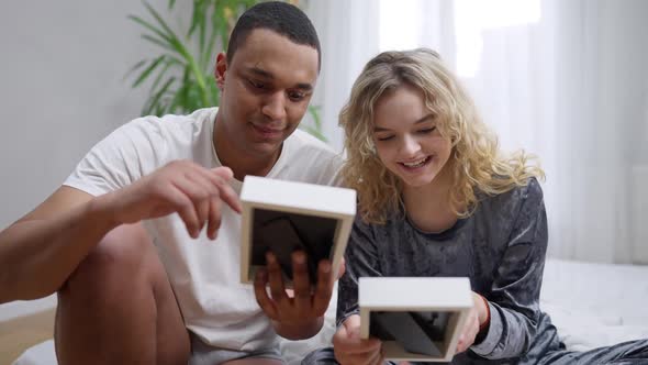 Happy Young Couple Talking Smiling Looking at Pictures Sitting in Bedroom in the Morning