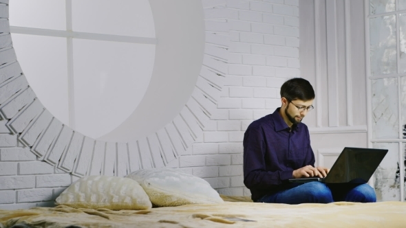 Bearded Man In Glasses Sitting On The Bed, Uses a Laptop