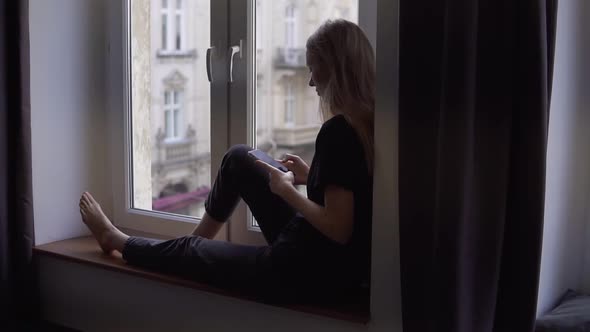 Rare View of a Woman Sitting on Window Sill and Using Smartphone at Home