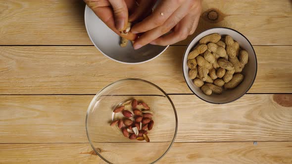 Viewed from above, men's hands peel peanuts from their shells, tossing the skins and nuts into diffe