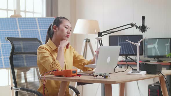 Asian Woman Sitting In Front Of Solar Cell Looking At Wind Turbine And Thinking While Working