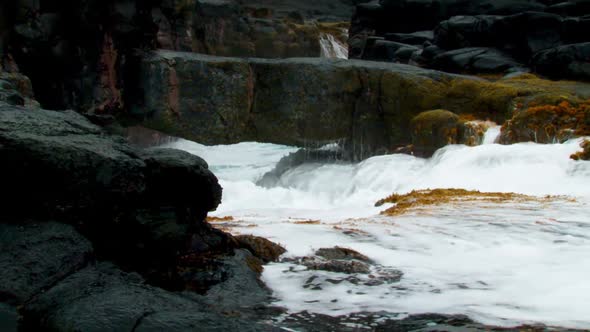 Rough ocean waves flow through a small channel and crash against moss-covered rocks along a rocky co