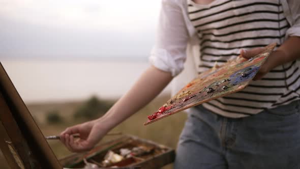 The Girl Artist in Striped Shirt Mixes the Colors on the Palette and Applies Them to the Canvas on