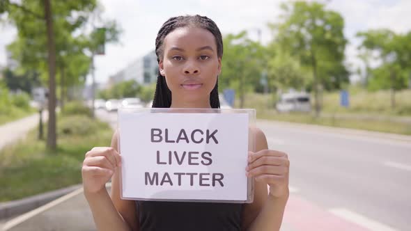 A Young Black Woman Holds Up a Black Lives Matter Sign in Front of the Camera - a Busy Road