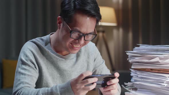 Close Up Of Asian Man Enjoys Playing Game On Smartphone After Working With Documents At Home