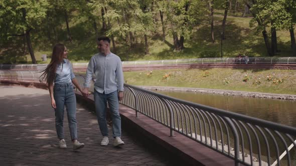 A Young Romantic Couple is Walking Along the Embankment in a City Park and Talking Holding Hands