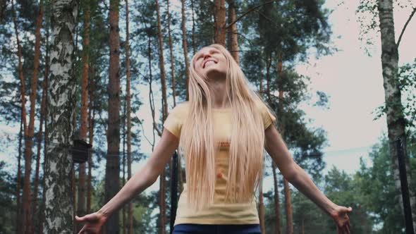 Happy Female Jumping on Trampoline. Beautiful Young Blond Barefoot Woman in Light Summer Shirt and