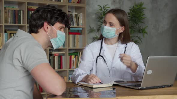 Woman Doctor Wearing a Medical Mask Sits at Her Desk and Chats to an Man Patient While Looking at