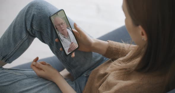 Female Medical Assistant Wears White Coat, Headset Video Calling Distant Patient on Smartphone